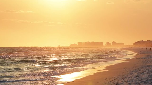 Dreamy pastel orange sunset in Santa Rosa Beach, Florida with Pensacola coast cityscape skyline, people walking in distance at panhandle with ocean waves washing on shore reflection