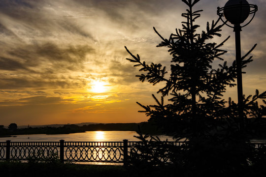contour image of the embankment of the river at sunset under a golden sky and clouds, railings and spruce