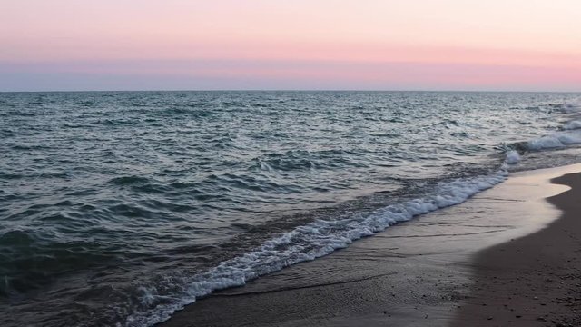 Waves break gently on a sandy Lake Michigan beach with a colorful sky illuminated by the pre-dawn rising sun. One minute loop.