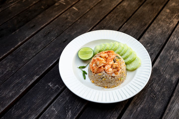 Thai style shrimps fried rice with fresh cucumber and lime on white ceramic plate on vintage wooden table background, healthy food