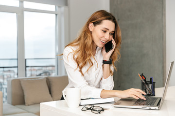 Obraz premium Business woman dressed in formal clothes shirt indoors talking by mobile phone using laptop computer.