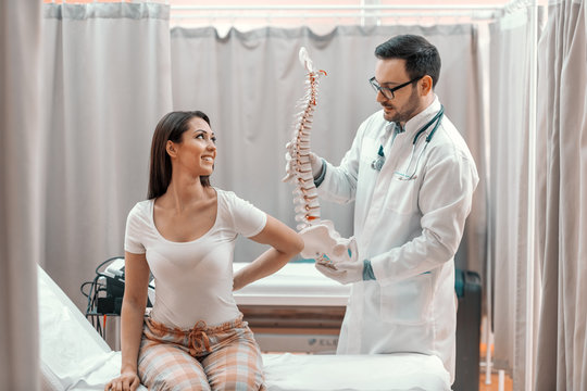 Female patient in pajamas sitting on hospital bed and showing hurting place on spine. Doctor standing next to her and holding spine model.
