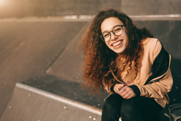 Smiling mixed race teenage hipster girl sitting at skate park and looking at camera.
