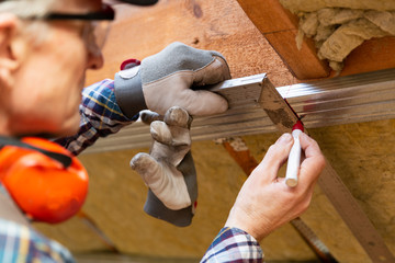 Man in googles making a mark on metal frame on unfinished attic ceiling. Attic insulation and...