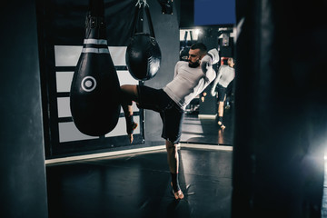 Muscular powerful Caucasian kick-boxer with boxing gloves on and in sportswear kicking bag in dark...