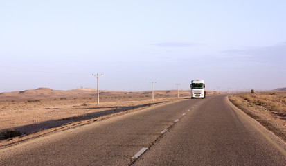 White truck goes to highway through scenic desert of Jordan. High voltage powerlines along asphalt road in arid valley. Early morning in wilderness after sunrise. Electric power poles. Horizontal.