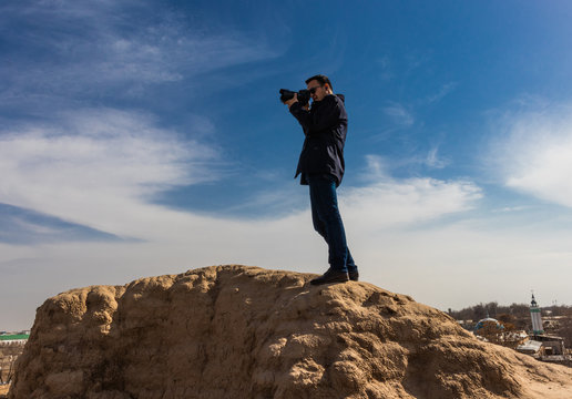photographer in dark outfit and black sunglasses holding a camera steady in his hands and making a shot while standing on top of an ancient wall. Aerial view.