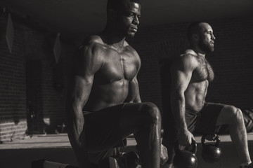 Fototapeta na wymiar Black and white photo of two young caucasian and african male athletes with kettlebells making lunges with weight in indoor workout
