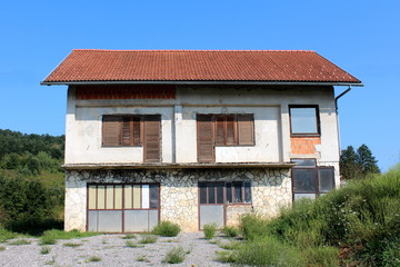 Fototapeta na wymiar Dilapidated abandoned old family house with ground floor turned into garage and closed doors and windows surrounded with high grass and gravel driveway in front and forest with clear blue sky in backg