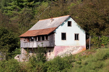 Obraz premium Dilapidated old family house with cracked facade and wooden balcony built on small hill surrounded with dense forest on warm summer day