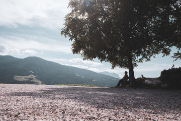 An elderly woman sits under a tree and enjoys the Alps panorama. Sunny view with some hazy sky