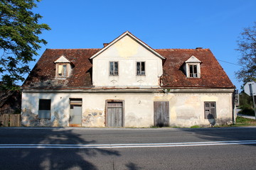 Elongated abandoned house with cracked facade and roof tiles broken windows and boarded doors next to paved road surrounded with tall trees and clear blue sky in background on warm sunny day
