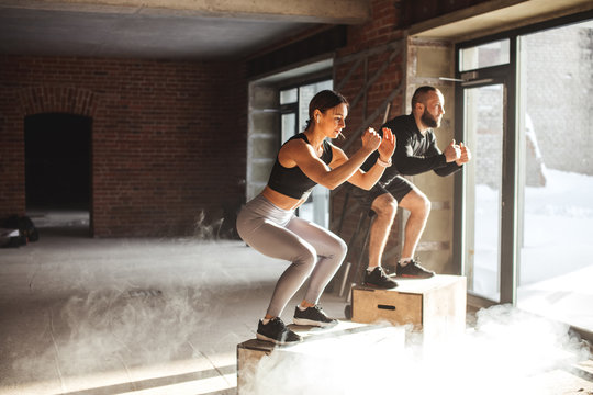 Caucasian sportive female and male athlets are performing box jumps at gym with dust and chalk particles floating in the air