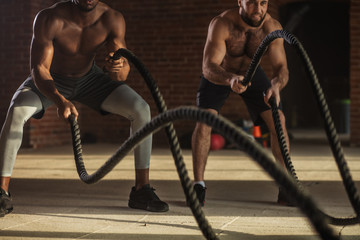 Muscular half-naked body athletes doing some crossfit exercises with a rope indoor, preparing to competitions and effectively burning excess fat.