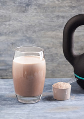Glass of Chocolate Protein Shake with milk and banana, Whey protein in scoop and black sporting kettlebell in background. Sport nutrition. Rustic wooden background. Copy space. 