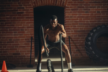 Fototapeta na wymiar Muscular powerful determined african man training with rope in functional training in outdoor gym with brick walls, strengthen the muscles of the shoulders, which is important for boxers and swimmers