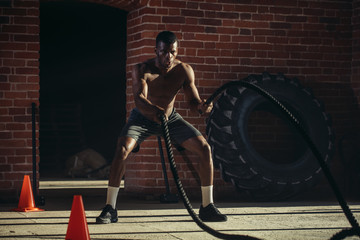 Handsome muscular bare chested gym man is doing battle rope exercise while working out at cross-fit training.