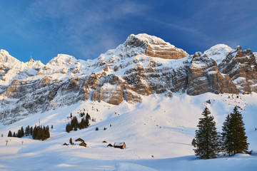 Schneebedeckte Schwägalp mit Alpsteinmassiv und Säntis im Winter, Felsen, blauer Himmel, leichte Schleierwolken, Häuser und Schäunen, Tannen