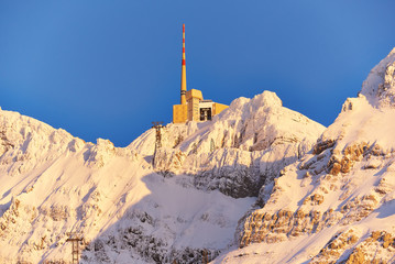 Säntis im Winter, Bergstation mit Sendemast, Nahaufnahme, schneebedecktes Felsmassiv im gelben Abendlicht, blauer Himmel, Seilbahnmasten