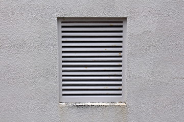 Grey metal protection bars on large industrial fan exit with partially rusted parts mounted on light grey wall on side of building on warm sunny day