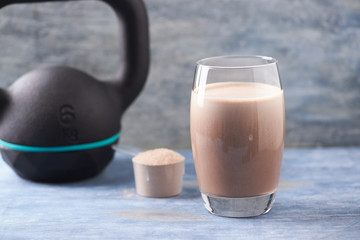 Glass of Chocolate Protein Shake with milk and banana, Whey protein in scoop and black sporting kettlebell in background. Sport nutrition. Rustic wooden background. Copy space. 