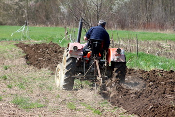 Man on older tractor plowing the field surrounded with uncut grass and dense trees in background on...