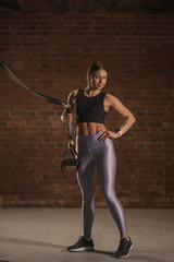 Portrait of athletic caucasian attractive fit woman dressed in black top and silver leggins, holding elastic TRX straps in hand posing against brick wall background. Sport, Fitness, Crossfit