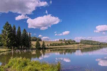 Fototapeta premium Summer view of the water surface of the river with overgrown picturesque banks and Cumulus clouds in the blue sky, toned.