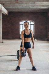 cheerful young sporty girl training with weight at gym. full length photo. happy sportswoman holding a kettlebell and looking at the camera