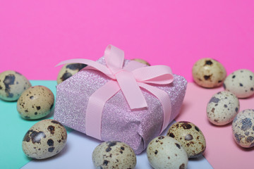 Quail eggs lie on the white surface. Next to the box with a gift wrapped in wrapping paper and tied with a ribbon. Sheets of colored paper for background.