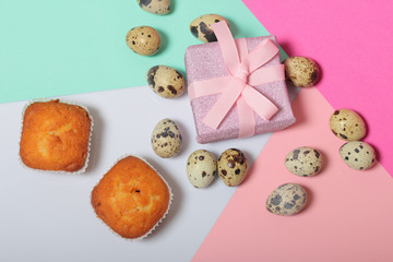 Quail eggs lie on the white surface. Nearby are cupcakes. Next to the box with a gift wrapped in wrapping paper and tied with a ribbon. Sheets of colored paper for background.