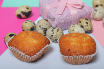 Quail eggs lie on the white surface. Nearby are cupcakes. Next to the box with a gift wrapped in wrapping paper and tied with a ribbon. Sheets of colored paper for background.