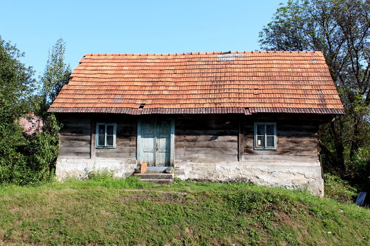 Small abandoned old wooden family house with dilapidated boards and broken roof with missing roof tiles on top of small hill surrounded with grass and trees with clear blue sky in background