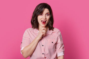 Portrait of happy pleased brunette woman with wavy hair, has pink makeup, licks lollipop, wears striped fashionable shirt, isolated over bright rosy background, being sweet tooth. Pinup girl indoor