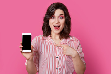 Fototapeta premium Portrait of independent, excited girl in pink, striped shirt with round buttons holding white mobile phone with black screen in fingers, and her second hand points to switched off smartphone.