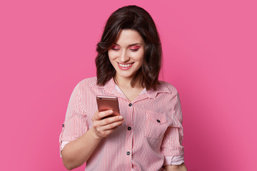 Сharming, beloved female person looks into smartphone with smile in middle of portrait photography...