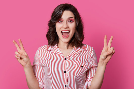 Optimistic fashionable woman with dark hair, smiles happily, makes peace gesture with both hands, dressed in stylish shirt, has fun indoor, isolated over pink background, has professional makeup