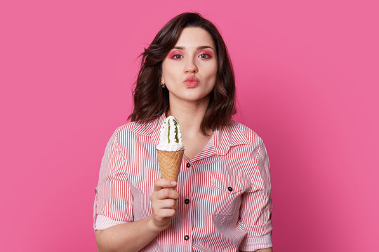 Headshot of attractive lady keeps lips folded, has pink makeup, eats tasty fresh ice cream, flirts with boyfriend, isolated over pink background, enjoys free time and summer vacation. Mmm. its yummy