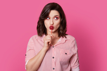 People, beauty, style concept. Good looking brown haired young woman with pink makeup, licks lollipop, keeps gaze aside, wears striped fashionable shirt, isolated over bright pink background