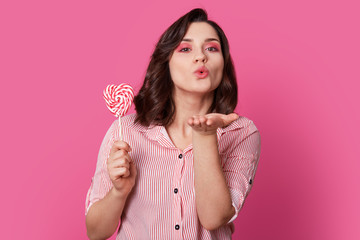 Photo of affectionate woman blows air kiss, dressed in fashionable clothes, holds candy, has bright makeup, poses over rosy background. Its love is for you. Lovely pinup girl has fun indoor.