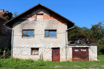 Suburban family brick house with attached garage and cracked damaged facade from shrapnels and bullets during war with boarded windows and heavily locked doors surrounded with uncut grass and forest v