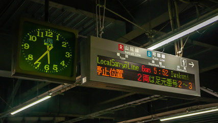 A huge rectangular electronic board showing the type of train line, departure time and destination in a typical train station in Japan, with a round analog clock.