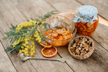 Composition of the candied orange spiral peel with sugar syrup in a glass jar and plate near the saucer with walnuts, mimosa, spoon and bread