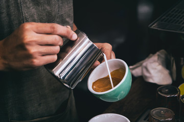 Close-up of male barista hand holding and pouring hot milk for prepare latte art on a green cup of cappuccino coffee. vintage color tone.