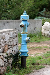 Fototapeta na wymiar Vintage rusted old blue metal fire hydrant mounted on strong metal pipe next to traditional stone wall surrounded with uncut grass and trees in background on warm sunny day
