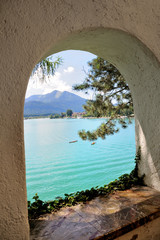 Beautiful mountain lake with turquoise water, the mountains in the background. Boats float on the lake. Sunny summer day, blue sky. View from the arched window of a stone building.