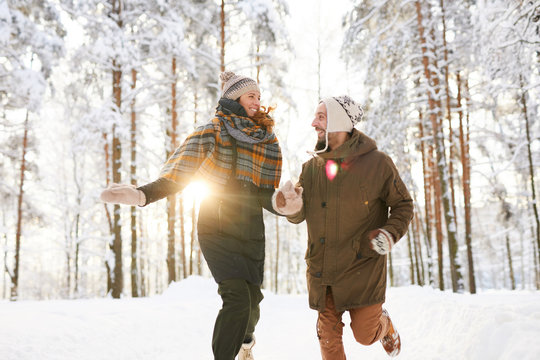 Portrait of happy playful couple running in winter forest and smiling happily in sunlight, copy space