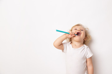 Caucasian little cute curly baby girl in white clothes body brushing her teeth with a colored blue brush on a white background.