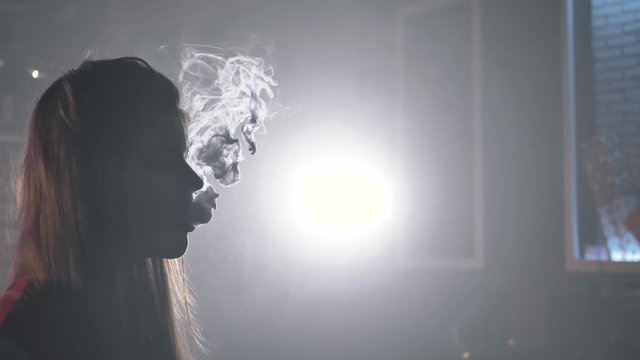 A medium shot of a backlit silhouette of a young girl making vape clouds