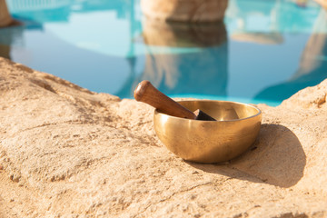 A singing Tibetan bowl stands on a sand-colored stone near a swimming pool on a sunny day.